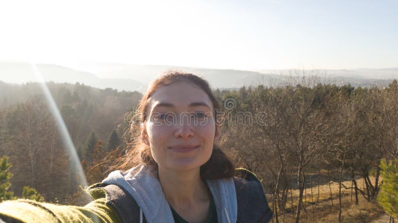 The woman smiles and takes a selfie from a hill on the background of the forest and mountains in the haze. A ray of sun stock photos The woman smiles and takes a selfie from a hill on the background of the forest and mountains in the haze. A ray of sun stock photos