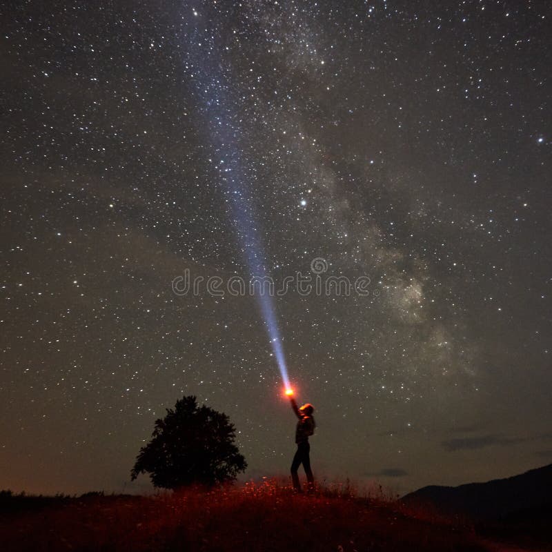 Woman standing against night starry sky with Milky Way in the mountains with flashlight in his hand