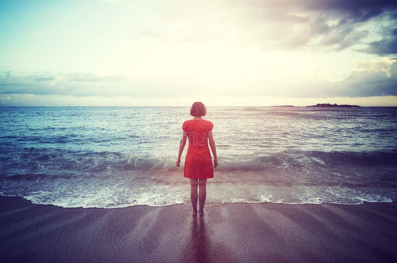 Woman Standing On A Beach Facing The Horizon At Sunset Stock Photo