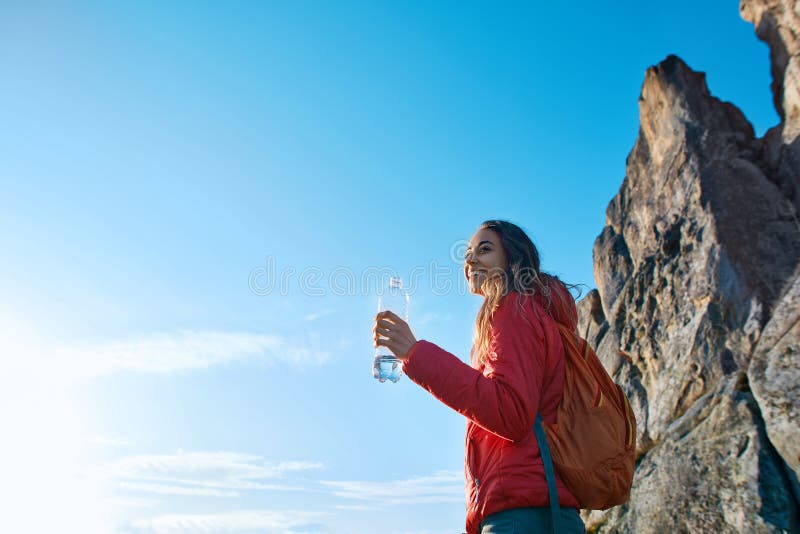 Woman hiker with backpack, wearing in red jacket, standing on edge of cliff against a blue sky background. Young female holds a bottle of water in hand and enjoying. Woman hiker with backpack, wearing in red jacket, standing on edge of cliff against a blue sky background. Young female holds a bottle of water in hand and enjoying.