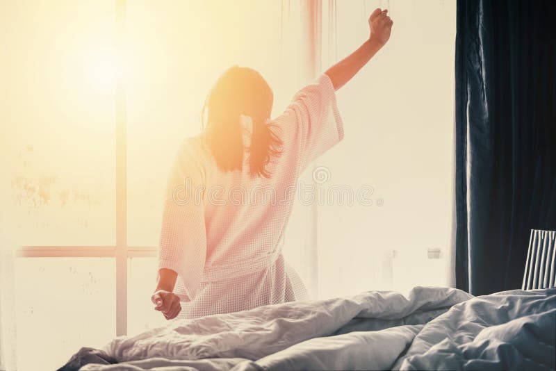 Woman stretching hands on bed after waking up while sitting on bed, entering a day happy and relaxed after good night sleep.