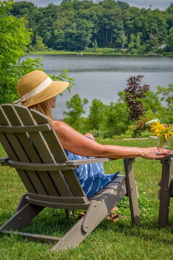 Woman In Sunhat Sitting In Adirondack Chair Along Lake 