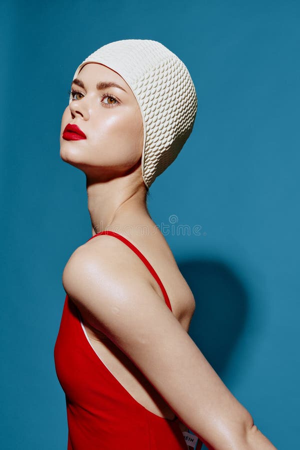 Woman swimmer in a red sexy swimsuit and red painted lips, wearing a white swim cap poses against a blue background