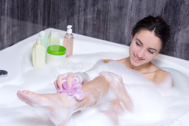 Beautiful smile woman washing leg with pink sponge in bath with foam, young...
