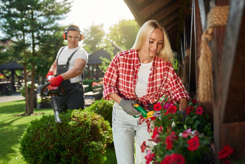 Beautiful woman in gloves working with gardening scissors near flowers while caucasian man trimming bushes on background. Two young people taking care of summer garden. Beautiful woman in gloves working with gardening scissors near flowers while caucasian man trimming bushes on background. Two young people taking care of summer garden.