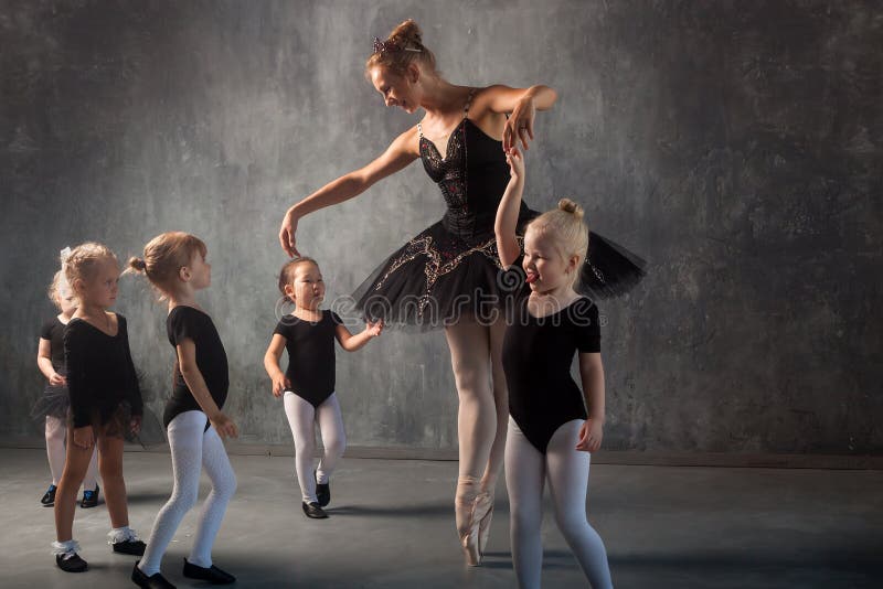 A beautiful blonde women in a black dance tutu, white pantyhose and pointe shoes is dancing and teaches young girls to dance ballet in a dark studio. A beautiful blonde women in a black dance tutu, white pantyhose and pointe shoes is dancing and teaches young girls to dance ballet in a dark studio