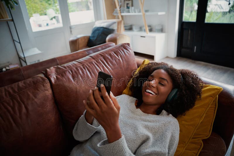 Woman with toothy smile lying on couch using smartphone at home listening to music on bluetooth wireless headphones stock images