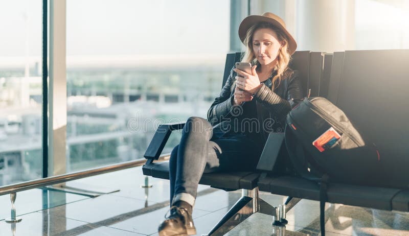 Woman tourist in hat, with backpack sits at airport near window, uses smartphone. Hipster girl waiting for plane landing royalty free stock photo Woman tourist in hat, with backpack sits at airport near window, uses smartphone. Hipster girl waiting for plane landing royalty free stock photo