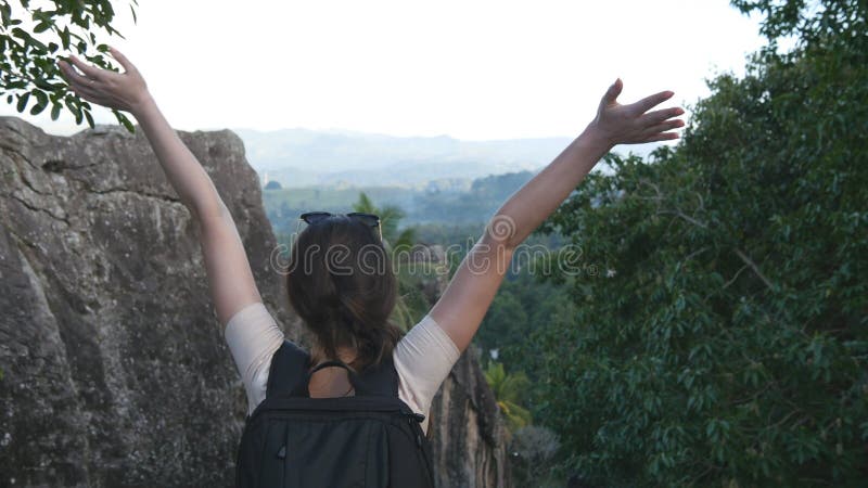 Woman tourist standing on the edge of beautiful canyon, victoriously outstretching arms up. Young female hiker with backpack reaching up top of mountain and raised hands. Rear back view.