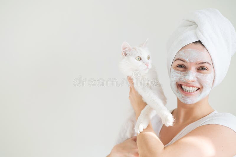 A woman with a towel on her hair and a clay mask on her face is holding a white fluffy cat on a white background. Copy