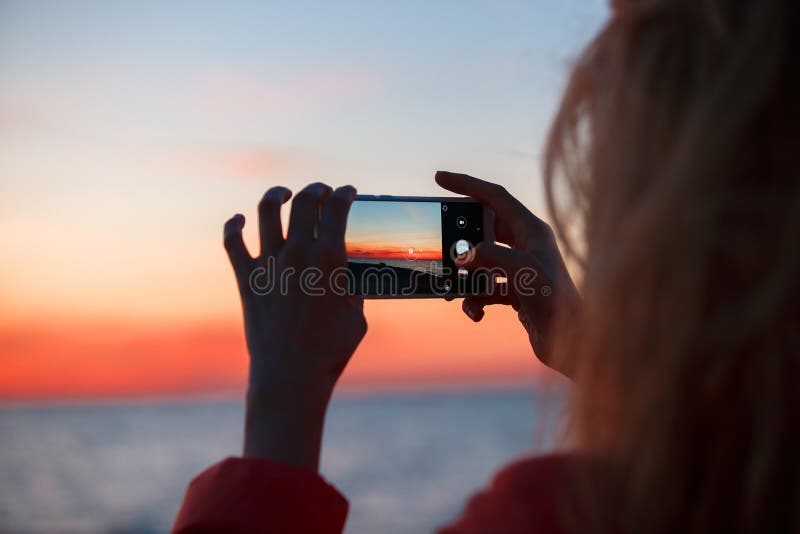 Woman traveler using smartphone and taking photo of colorful sea sunset