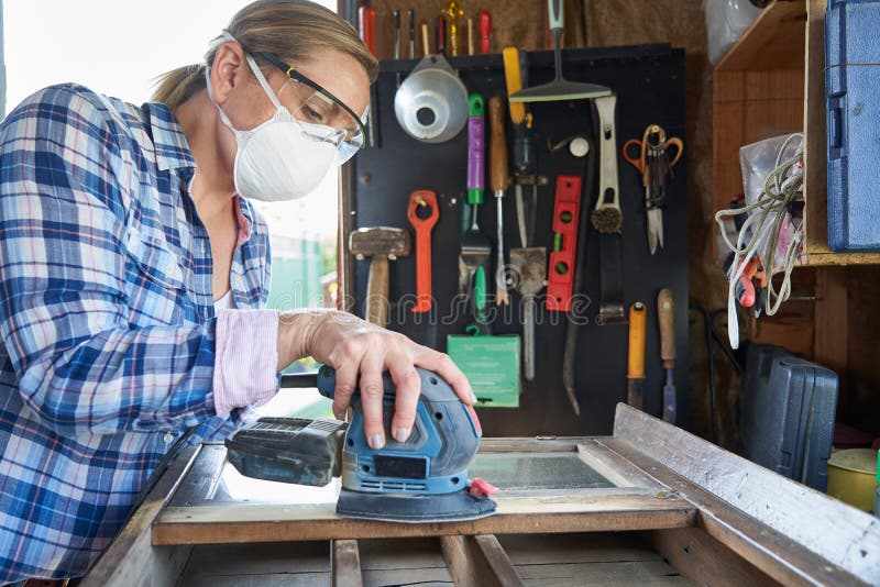 Mature Woman Upcycling Furniture In Workshop At Home Using Electric Sander