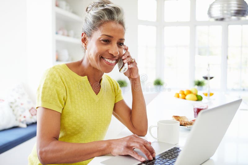 Woman Using Laptop And Talking On Phone In Kitchen At Home