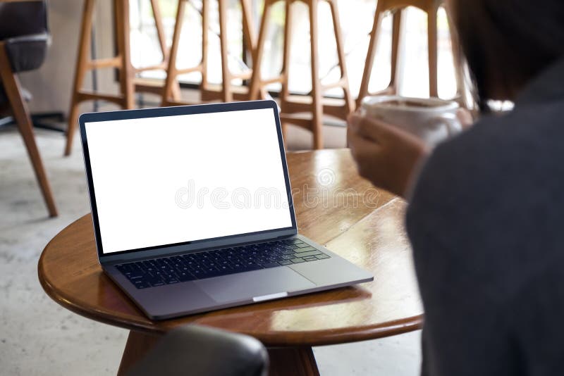 A woman using and looking at laptop with blank white desktop screen while drinking hot coffee in cafe