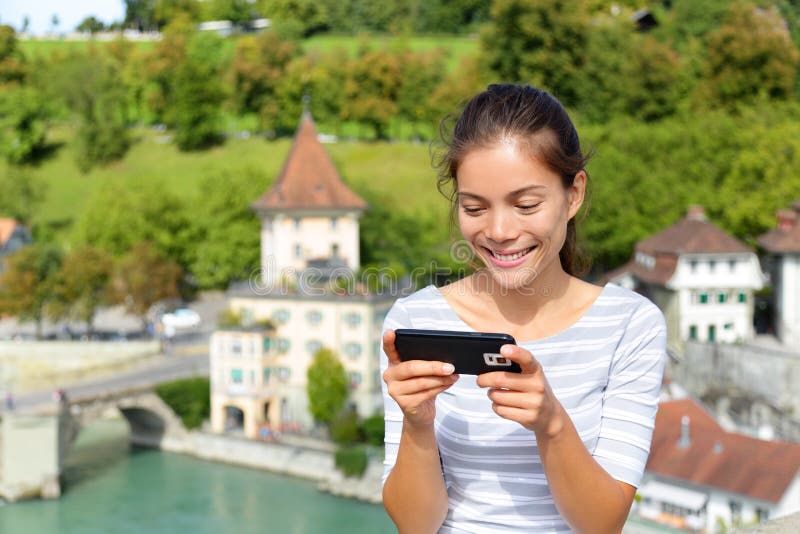 Woman using smart phone app in Bern Switzerland, Europe. Person using smartphone application on travel. Happy smiling Asian girl sitting on Nydegg bridge by the Aare river in the Swiss city of Berne