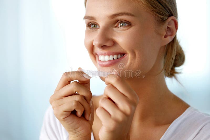 White Smile. Beautiful Smiling Woman With Healthy White Teeth Applying Teeth Whitening Strip. Portrait Of Happy Girl Using Dental Whitener. Dental Beauty, Tooth Care Concept. High Resolution Image. White Smile. Beautiful Smiling Woman With Healthy White Teeth Applying Teeth Whitening Strip. Portrait Of Happy Girl Using Dental Whitener. Dental Beauty, Tooth Care Concept. High Resolution Image