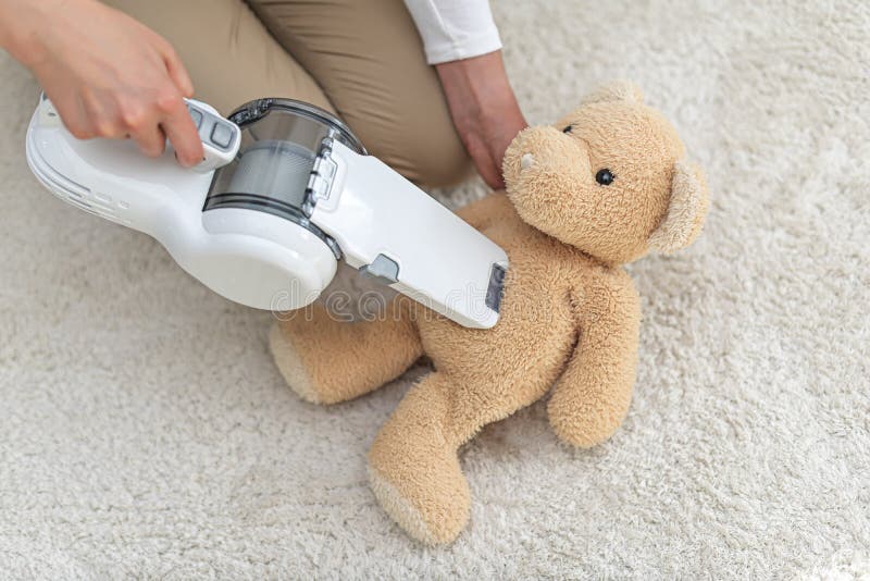 Woman vacuuming Teddy bear in a house with a hand-held portable vacuum cleaner.