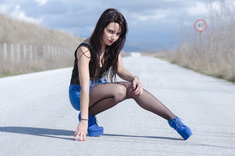 Woman in profile wear a blue skirt and black stockings sitting on legs on the open road, cloudy sky as background, horizontal and full length photo. Woman in profile wear a blue skirt and black stockings sitting on legs on the open road, cloudy sky as background, horizontal and full length photo