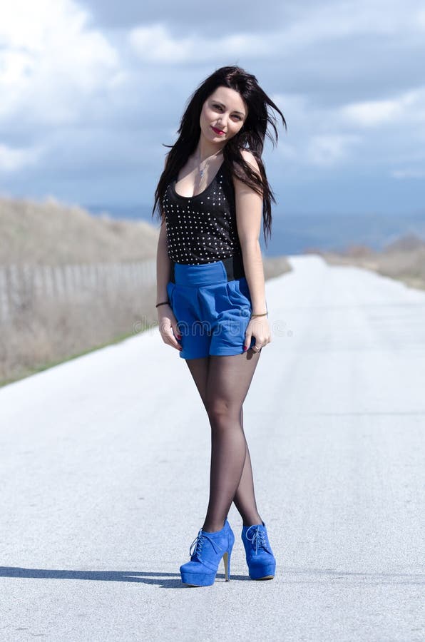 Woman wear a blue skirt and black stockings standing upright with crossed legs in the middle of empty road, cloudy sky as background, vertical and full length photo. Woman wear a blue skirt and black stockings standing upright with crossed legs in the middle of empty road, cloudy sky as background, vertical and full length photo