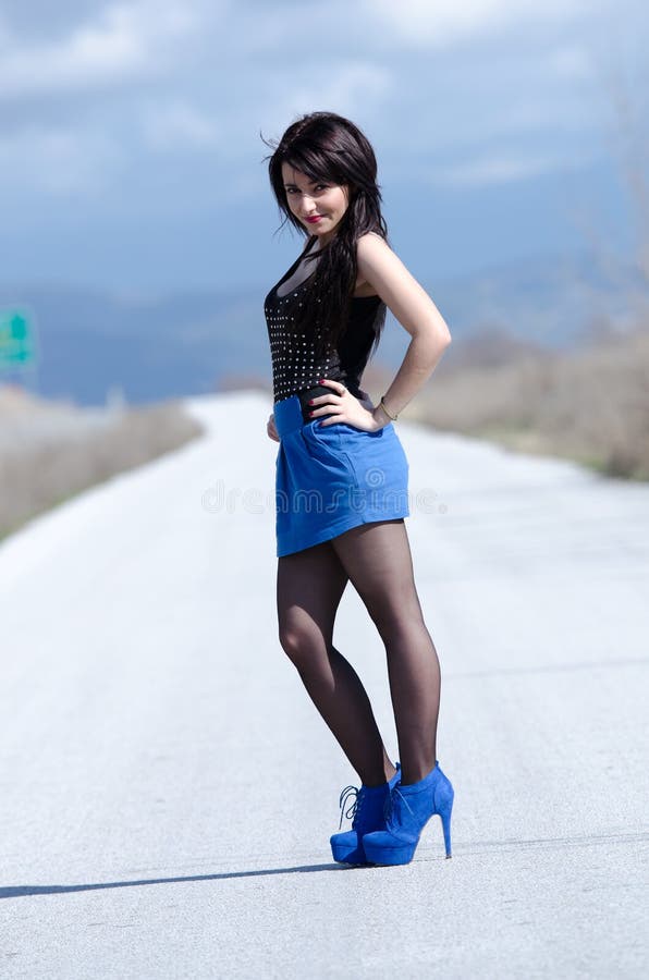 Woman wear a blue skirt and black stockings standing upright in profile in the middle of empty road, cloudy sky as background, vertical and full length photo. Woman wear a blue skirt and black stockings standing upright in profile in the middle of empty road, cloudy sky as background, vertical and full length photo