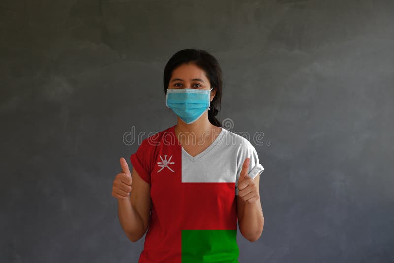 Woman wearing hygienic mask and wearing Oman flag colored shirt with thumbs up with both hands on dark wall background stock images