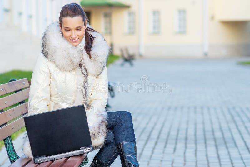 Young smiling beautiful woman in white coat with fur collar sitting on bench and holding laptop on your lap. Young smiling beautiful woman in white coat with fur collar sitting on bench and holding laptop on your lap.