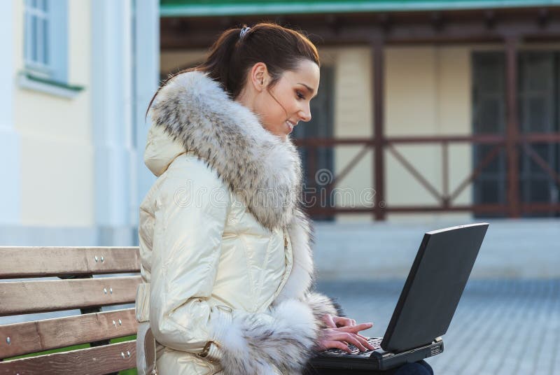 Young smiling beautiful woman in white coat with fur collar sitting on bench and holding laptop on your lap. Young smiling beautiful woman in white coat with fur collar sitting on bench and holding laptop on your lap.