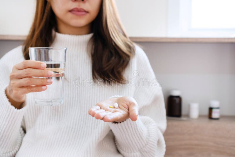 Woman in white sweater holding pill and glass of water in hands taking emergency medicine