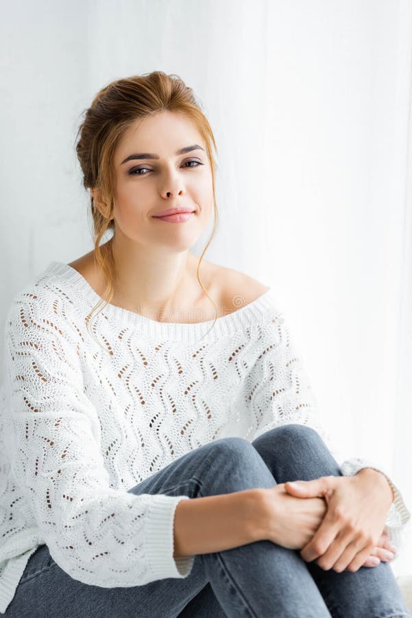 Woman In White Sweater And Jeans Sitting And Looking At Camera Stock