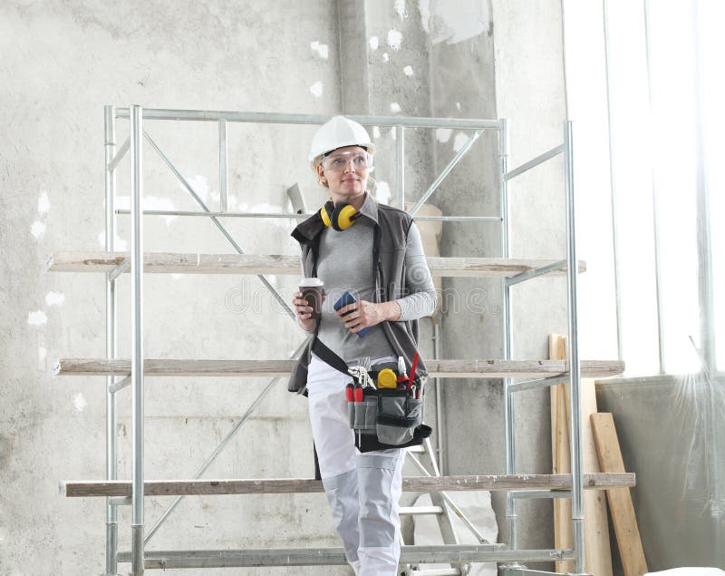 Woman worker builder to coffee break with mobile phone, wearing helmet, glasses, bag tools and ear protection headphones royalty free stock images