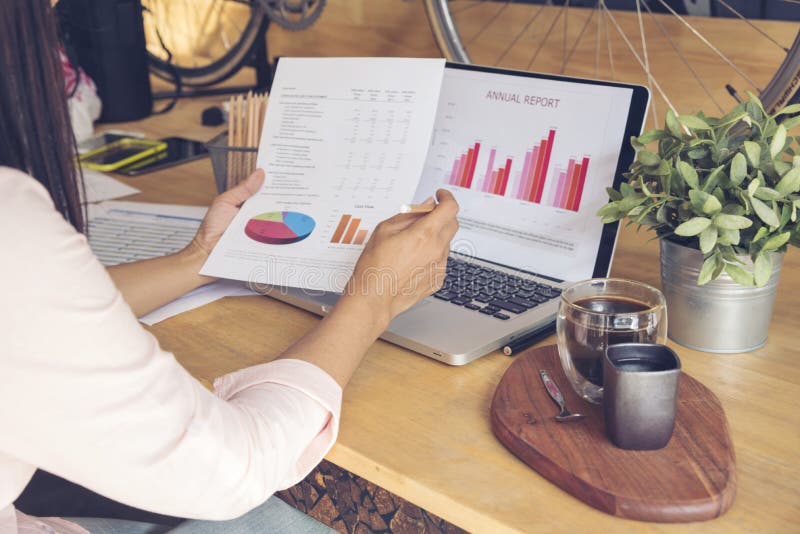 Woman working at home office desk using laptop business financial document chart and graph on wooden table with coffee cup. Woman