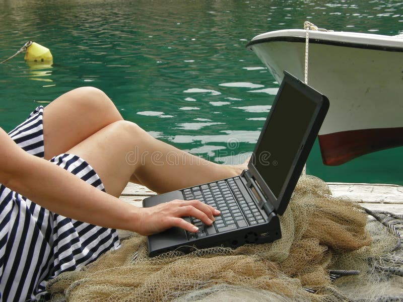 View at a attractive woman legs (mariner) work on laptop in harbor with boat, beacon and fishing net. Horizontal color photo. View at a attractive woman legs (mariner) work on laptop in harbor with boat, beacon and fishing net. Horizontal color photo.