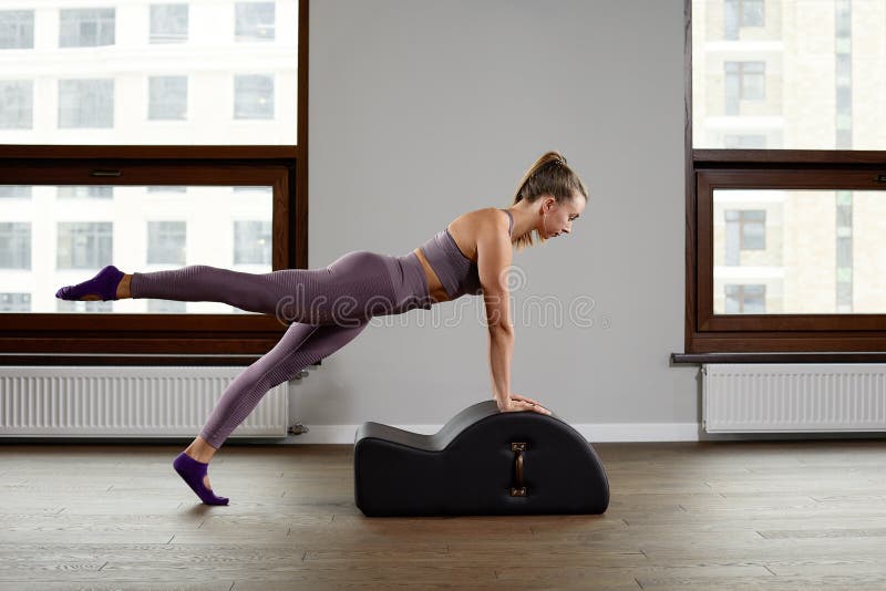 A woman yoga instructor trains on a reformed cadilac in a large hall against a window, modern equipment for training in