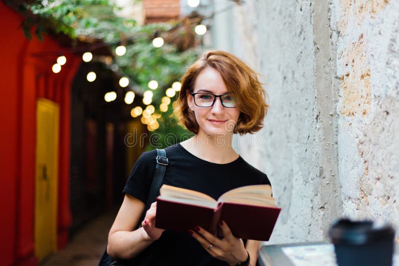 Young hipster woman in glasses with short haircuts reading book in outdoor street cafe and looking at camera, attractive, casual, female, lifestyle, happy, literature, leisure, outdoors, studying, girl, portrait, education, sunshine, beautiful, knowledge, relaxation, recreation, indoor, adult, city, summer, intellectual, people, novel, beauty, caucasian, smiling, cheerful, clothing, student, learning, holding, pretty, garlands