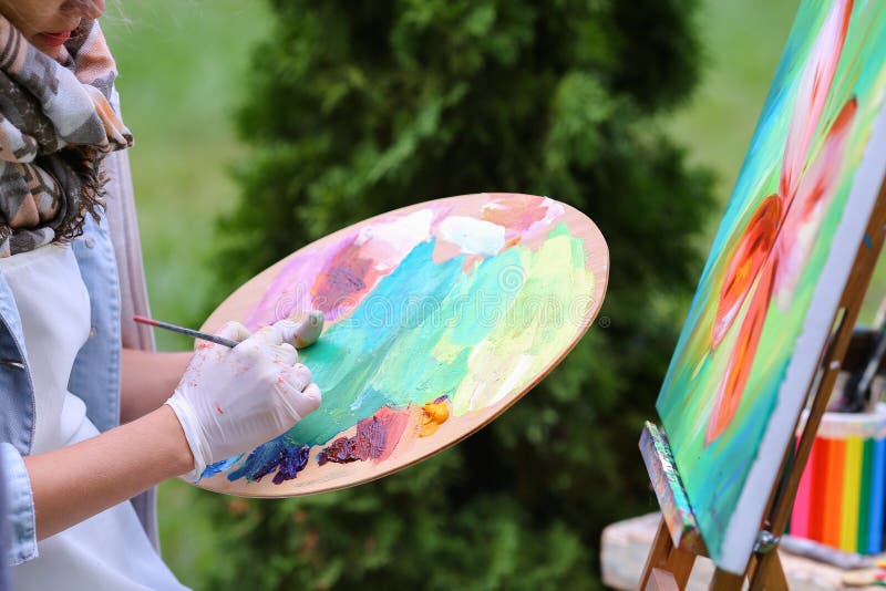 Photo of female hand artist who mixes oil or acrylic colored bright colors on wooden panel, which would start to draw and located in green park in open air. Concept of set of objects to paint, oil or acrylic paints, advertising art school. Photo of female hand artist who mixes oil or acrylic colored bright colors on wooden panel, which would start to draw and located in green park in open air. Concept of set of objects to paint, oil or acrylic paints, advertising art school.