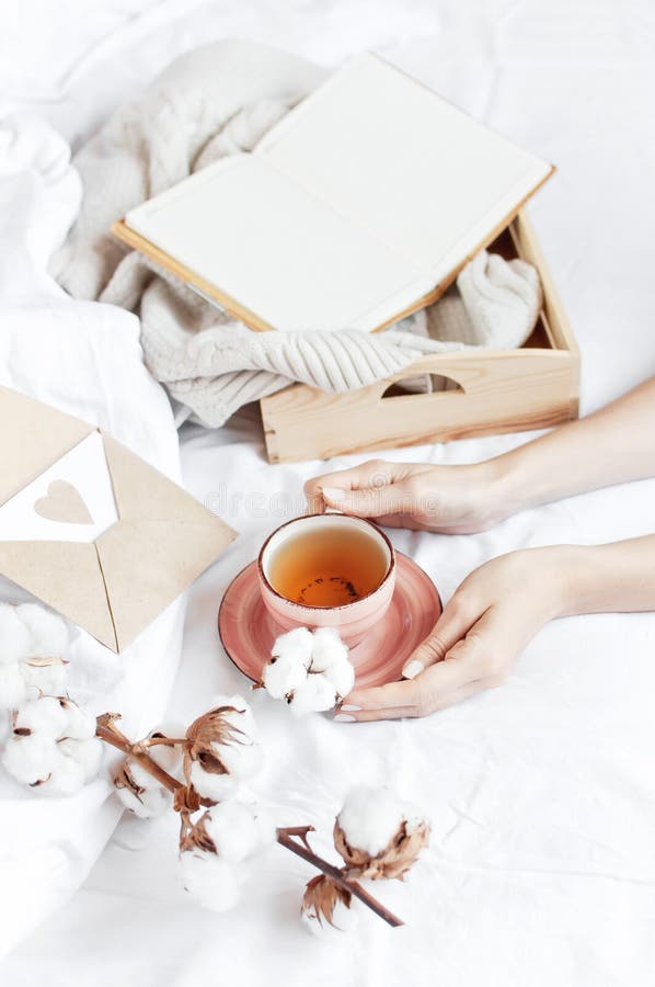 Women hands hold cup of hot tea with delicate cotton flower open notebook love letter tray white knitted sweater in the bed. Cozy