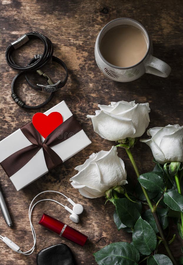 Women`s table. Bouquet of roses, gift box, leather bracelets, lipstick, headphones, coffee on a dark wooden background, top view. stock photos Women`s table. Bouquet of roses, gift box, leather bracelets, lipstick, headphones, coffee on a dark wooden background, top view. stock photos