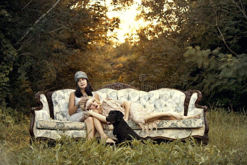 Bionda e bruna donne in vent'anni di moda, posando su di un vintage Vittoriano divano all'aperto con tramonto e un sacco di verde.