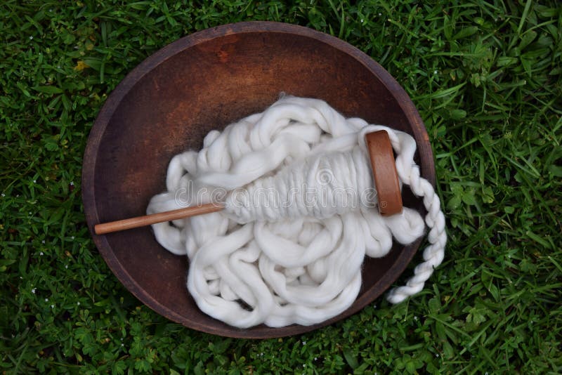 This is a wooden bowl filled with natural white cotton pencil roving with a wood spindle filled with cotton yarn and a spinning wheel bobbin filled with yarn on a green grass background. This is a wooden bowl filled with natural white cotton pencil roving with a wood spindle filled with cotton yarn and a spinning wheel bobbin filled with yarn on a green grass background.