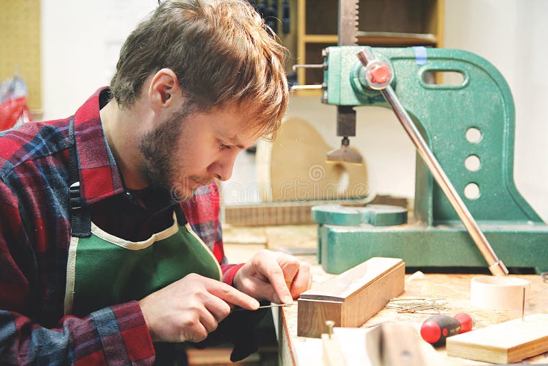 A young man who works as a luthier is building a wood acoustic guitar in his home workshop. A young man who works as a luthier is building a wood acoustic guitar in his home workshop.
