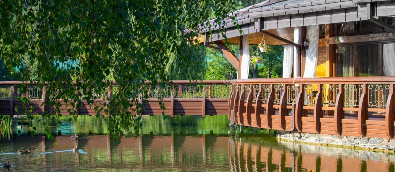 Wooden alcove near lake with ducks and trees. Garden house with reflection in water.