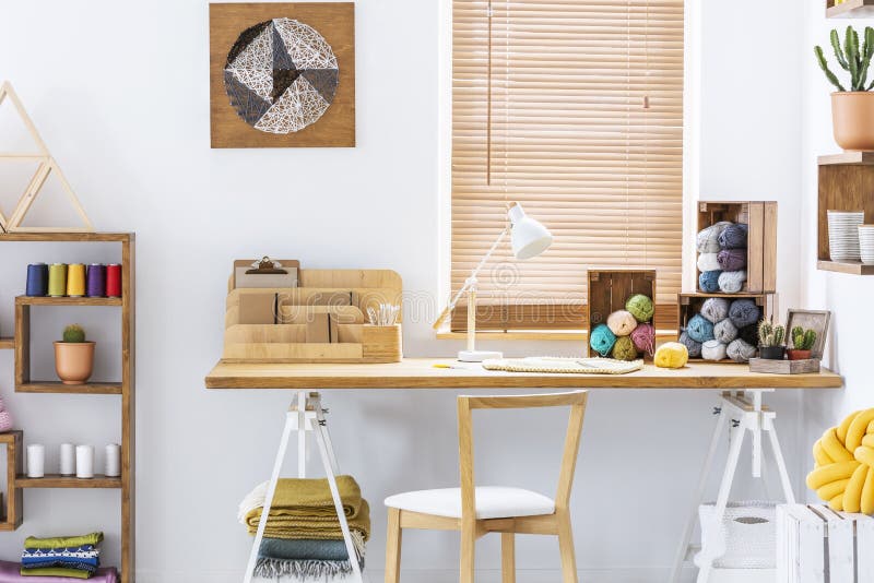 Wooden chair at desk with lamp and colorful yarns in home office interior with poster. Real photo