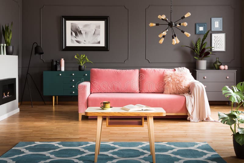 Wooden coffee table with tea cup and open book standing on carpet in dark living room interior with powder pink lounge with fur cushion and blanket concept