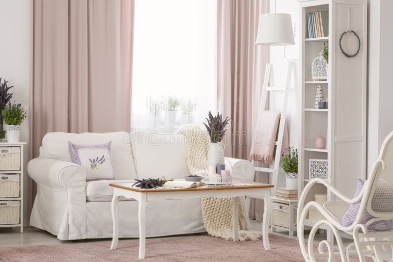 Wooden table next to white couch in pink living room interior with drapes and plants. Real photo concept