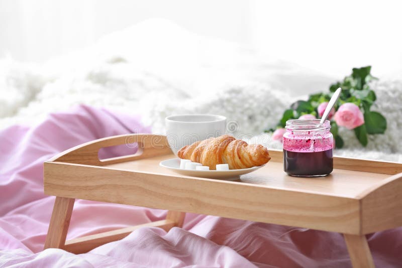 Wooden tray with tasty croissant, cup of coffee and jam on bed