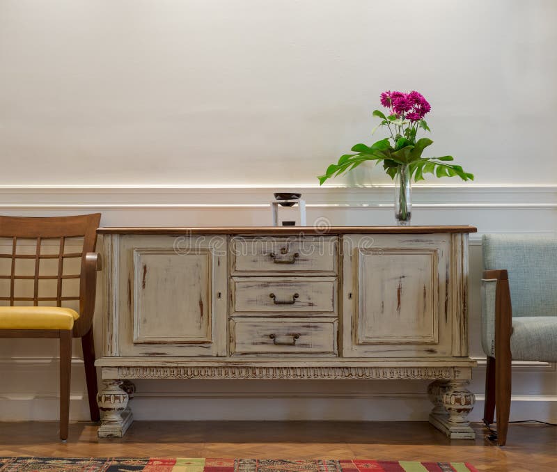 Wooden vintage off white sideboard, glass vase with red flowers, two chairs on white wall and wooden parquet floor