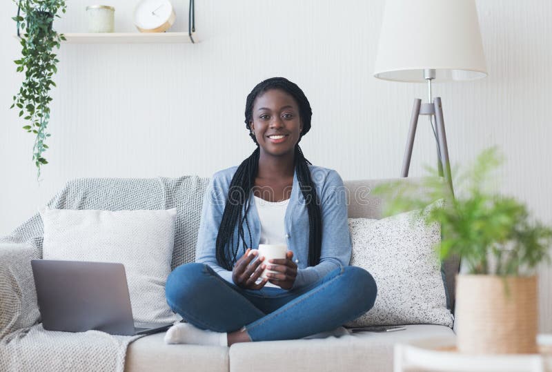 Work from home. Portrait of smiling black woman freelancer sitting on couch with coffee and laptop. Work from home. Portrait of smiling black woman freelancer sitting on couch with coffee and laptop