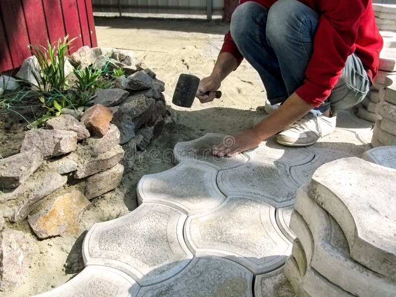 A worker beats a surface of the gray figured paving slab with a mallet, setting it in a dry sand-cement mix. Concept of the stock photography