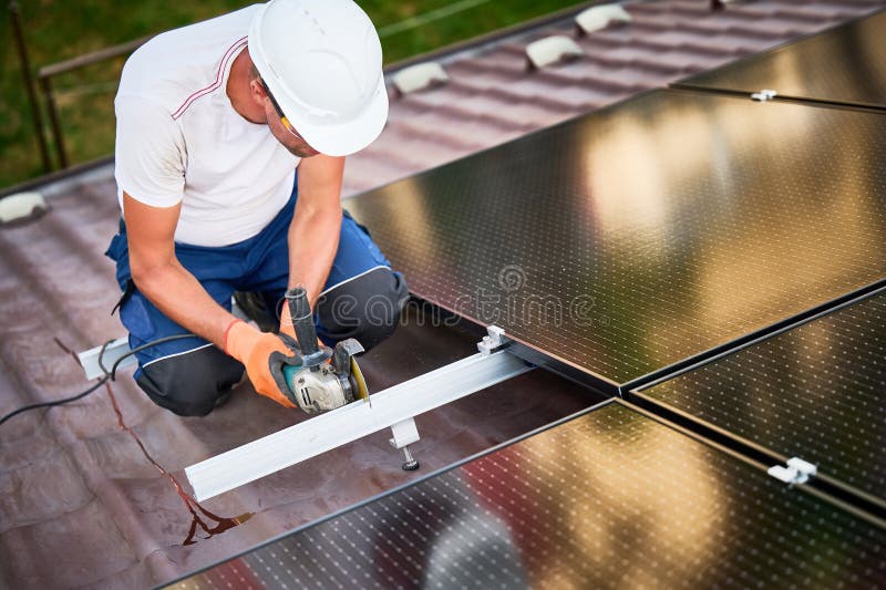 Worker installing solar panel system on roof using grinder on metal part of panel mounting structure. Worker installing or maintaining photovoltaic solar panel system on roof of house. Technician in helmet using grinder on metal part of the panel& x27;s mounting structure. Concept renewable energy.