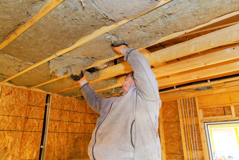 Construction worker installing insulation in residential building under wooden beams during interior renovation stock photography